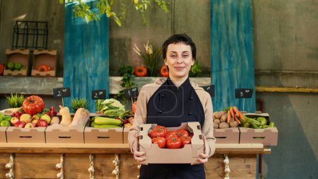 Foto de Mujer agricultora sosteniendo caja de tomates bio frescos en el mercado de alimentos saludables, la venta de productos de cosecha propia. Joven vendedor feliz presentando frutas y verduras naturales orgánicas en el mercado de agricultores. - Imagen libre de derechos