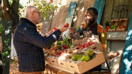 Téléchargez les photos : Group of diverse people talking about farmers market products on stand, looking at healthy products on counter. Elderly man and local stall holder choosing organic fruits and veggies. - en image libre de droit