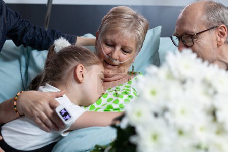 Téléchargez les photos : Little girl waving goodbye to sick grandmother. Elderly woman lying in hospital bed connected to medical monitoring devices hugging granddaughter. Relatives visiting old ill lady in geriatric clinic. - en image libre de droit