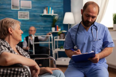 Foto de Male medical assistant wearing uniform and stethoscope filling out medical history of senior patient in wheelchair taking notes on clipboard. Woman providing personal information to nurse. - Imagen libre de derechos
