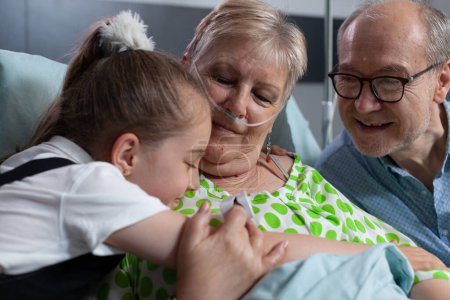 Elderly couple being visited by little girl at nursing home room. Smiling granddaughter showing love to happy grandparents, hugging at medical clinic room. Close up of hug between toddler, old lady.