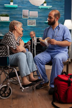 Foto de Male nurse giving patient support by reading stories from books to elderly woman with disability in living room. Nursing social services to senior people in health center. - Imagen libre de derechos