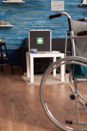 Photo for Wheelchair in nursing home with first aid case on waiting room table in background. Common area without people in it, available for patients in need of physical therapy and rehabilitation sessions. - Royalty Free Image