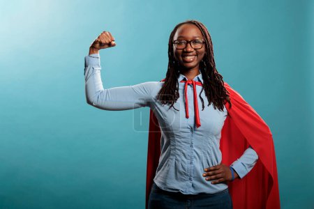 Photo for Arms muscle flexing brave superhero woman posing strong and tough for camera on blue background. Justice defender wearing mighty hero red cape while expressing empowerement. Studio shot. - Royalty Free Image