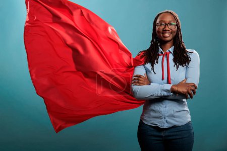 Foto de Mighty looking tough superhero woman wearing red hero costume cape standing with arms crossed on blue background. Portrait of young adult person posing as justice defender while smiling at camera. - Imagen libre de derechos
