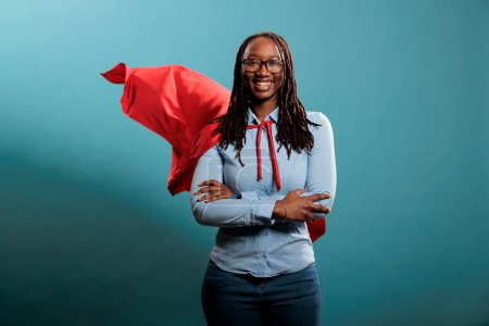 Photo for Confident looking mighty powerful young adult person wearing superhero costume on blue background. Portrait of happy brave woman with superpowers wearing red hero cape while standing with arms crossed - Royalty Free Image