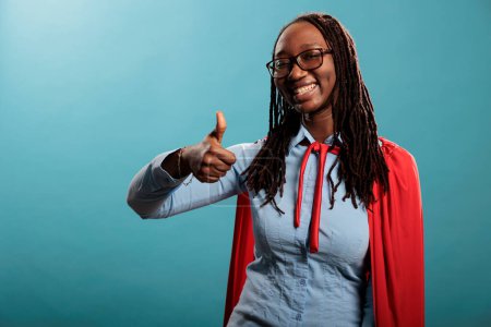 Photo for Young justice defender wearing red hero mighty cape showing thumbs up gesture sign at camera on blue background. Brave and proud superhero woman wearing cloak while smiling heartily. Studio shot. - Royalty Free Image