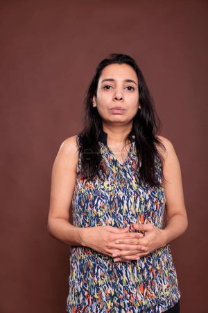 Foto de Indian woman with neutral facial expression, intertwined fingers portrait. Calm lady, young person standing with serious face front view studio medium shot on brown background - Imagen libre de derechos