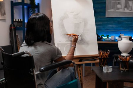 Téléchargez les photos : African american female professional artist using wheelchair to get around performing artwork on canvas easel in home drawing studio. Young woman with artistic skills making illustration at workplace. - en image libre de droit