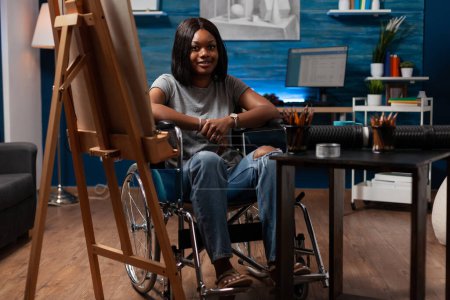Téléchargez les photos : Young african american female professional artist wheelchair user looking at camera from home art studio. Portrait of woman cartoonist surrounded by creative tools in atelier at home. - en image libre de droit