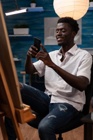 Foto de Smiling young man sharing on social media photo of finished artistic sketch with smart phone. African american art student showing canvas to classmates through video call with cell phone. - Imagen libre de derechos