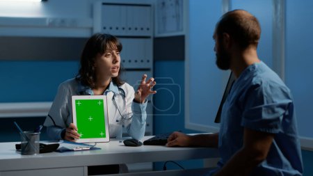 Foto de Medic holding tablet computer pointing at green screen chroma key display while discussing patient report with assistant. Clinical staff working night shift at medical expertise in hospital office - Imagen libre de derechos
