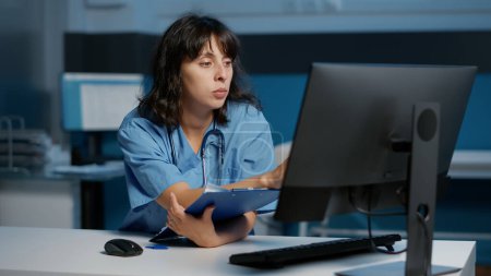Photo for Physician nurse checking illness report on computer while working at patient treatment to help cure disease during night shift in hospital office. Medical assistant looking at papers with expertise - Royalty Free Image