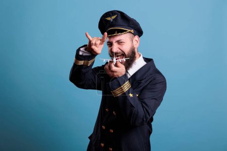 Téléchargez les photos : Funny aviator in aviation uniform playing with small airplane toy, academy captain holding commercial passenger plane model. Aircraft crew member looking at camera, studio medium shot - en image libre de droit
