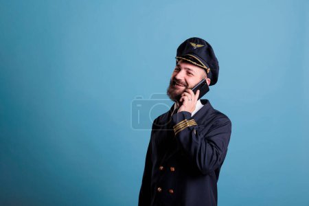 Foto de Middle aged captain talking on modern phone, holding smartphone, having conversation. Airlane aviator in uniform with calm neutral facial expression answering telephone call - Imagen libre de derechos