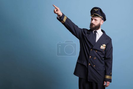 Photo for Airplane pilot pointing at sky with index finger, wearing professional uniform, plane captain looking at camera. Confident aviation academy aviator standing, studio medium shot - Royalty Free Image