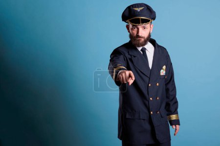 Photo for Serious aircraft pilot pointing at camera, plane aviator wearing uniform and hat front view portrait. Aviation academy captain with airline wings badge on jacket looking at you - Royalty Free Image