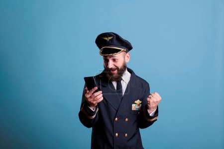 Photo for Happy excited airplane captain in uniform holding smartphone with winner gesture, clenched fist. Pilot with cheerful facial expression looking at mobile phone screen, reading good news - Royalty Free Image