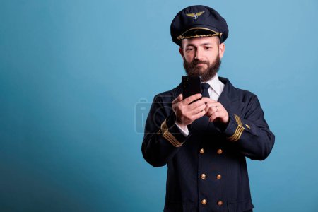 Photo for Portrait of plane capitan holding smartphone texting with remote aircrew member in studio with blue background. Pilot with aviation uniform, watching online video on mobile phone, medium shot - Royalty Free Image