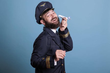 Photo for Pilot in uniform playing with airplane model side view, aviation academy aviator holding commercial plane toy. Funny aircraft crew member, studio medium shot on blue background - Royalty Free Image