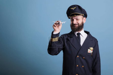 Foto de Smiling pilot in uniform holding small airplane model front view, aviation academy aviator playing with commercial plane toy. Aircraft crew member looking at camera, studio medium shot - Imagen libre de derechos