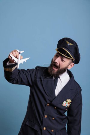 Photo for Pilot in uniform playing with airplane model, aviation academy aviator demonstrating plane landing training. Aircraft crew member holding commercial jet toy, studio medium shot - Royalty Free Image