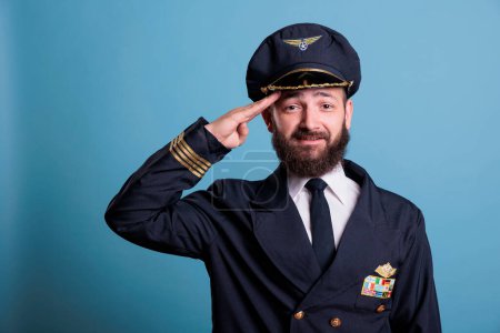 Photo for Smiling airplane captain saluting, wearing aviation uniform and hat front view portrait, plane captain looking at camera. Aviation academy pilot with airline wings badge on jacket - Royalty Free Image