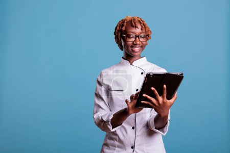 Photo for Female chef with digital tablet standing in studio while looking up recipes for new menu items. Head chef with mobile device providing garnish ideas for a gourmet cuisine meal. - Royalty Free Image