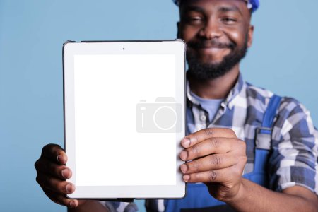 Téléchargez les photos : Smiling construction worker holding digital tablet with blank screen for advertising in front of camera in studio shot with blue background.Builder showing electronic device with copy space. - en image libre de droit
