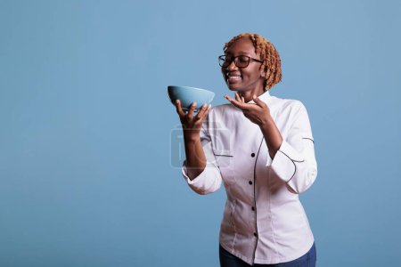 Téléchargez les photos : African american female chef holding blue bowl while delighting in aroma of new recipe she has just created. Cook dressed in work uniform posing in studio shot against blue background. - en image libre de droit