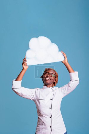 Téléchargez les photos : Cheerful woman wearing chef jacket holding cloud of text above head with space for advertising. Smiling cook with white text bubble in studio shot against blue background. - en image libre de droit