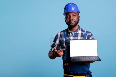 Téléchargez les photos : Cheerful builder pointing at empty laptop display in studio, advertising copy space accessory for text message. Handyman wearing helmet and tools belt showing blank screen advertising. - en image libre de droit