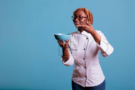 Foto de Female african american cook proudly showing light blue bowl of food, posing in studio. Relaxed professional female chef wearing work uniform and glasses against blue background. - Imagen libre de derechos