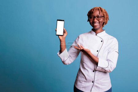 Photo for Young curly haired, kitchen chef with glasses, happily showing recipes on smartphone screen. Young African american woman, professional cooker holding portable cell phone. - Royalty Free Image