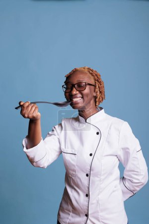 Téléchargez les photos : Smiling cooking professional wearing chef jacket testing new recipe with spoon in studio shot. Optimistic african american chef in uniform excited about new dish preparation against blue background. - en image libre de droit