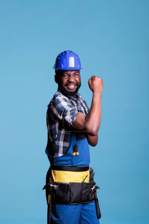 Photo for Strong builder flexing biceps and triceps muscles on camera, showing masculinity. Construction worker used to heavy work proud of strength, posing against blue background in studio. - Royalty Free Image