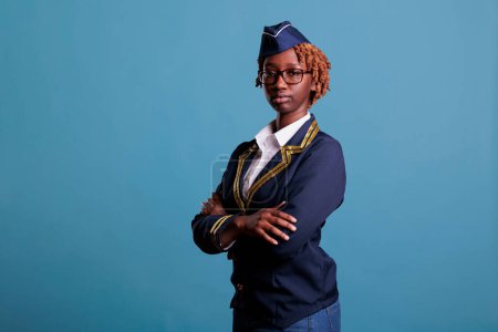Téléchargez les photos : Close-up view of a flight attendant with arms crossed, tired from all the work in a studio shot. Stewardess in uniform and glasses appearing serious while looking at camera against blue background. - en image libre de droit