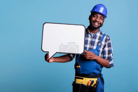 Téléchargez les photos : Cheerful construction employee holding blank template advertisement poster. Confident and smiling african american male holding white cardboard speech bubble on blue background. - en image libre de droit