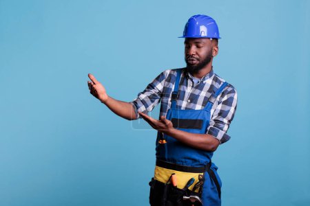 Foto de Construction worker wearing hard hat pretending to hold advertising banner in studio shot against blue background. Builder with arms raised as if carrying something and looking intently. - Imagen libre de derechos