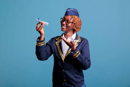 Foto de African american flight attendant in work uniform holding a model airplane. Stewardess showing commercial scale airplane with space for airline advertising in studio shot. - Imagen libre de derechos