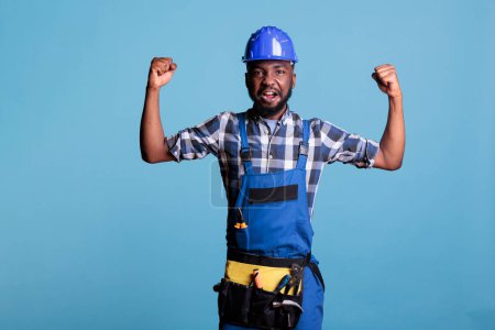 Photo for African american construction worker in coveralls showing his arm muscles in front of the camera. Construction worker in hard hat demonstrating his strength against blue background in studio shot. - Royalty Free Image