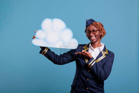 Photo for Female flight attendant smiling while holding paper cloud against blue background in studio shot. Optimistic woman stewardess wearing uniform, concept of flying among the clouds. - Royalty Free Image