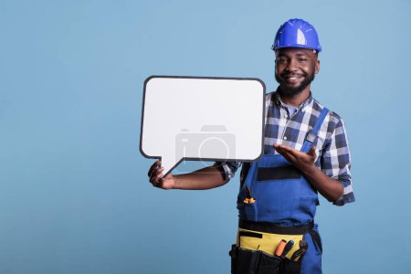 Foto de Smiling construction worker pointing to blank dialogue cloud with copy space, advertising mockup. Man holding speech bubble with message frame looking at camera, studio shot against blue background. - Imagen libre de derechos