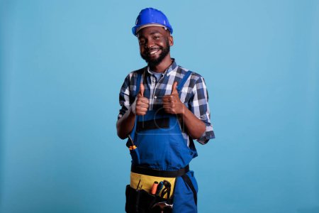 Photo for Studio portrait of proud electrician giving thumbs up in approval. Optimistic construction worker wearing work uniform hard hat and tool belt isolated on blue background. - Royalty Free Image