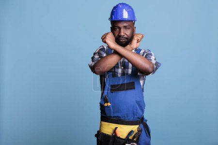 Photo for Disapproving african american builder making stop gesture with crossed arms while standing on blue background. Construction worker showing deny hands symbol while refusing to accept more work. - Royalty Free Image