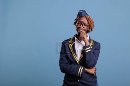 Photo for African american female flight attendant with doubtful expression wearing uniform against blue background in studio shot. Flight attendant thinking about the best in flight service strategy. - Royalty Free Image