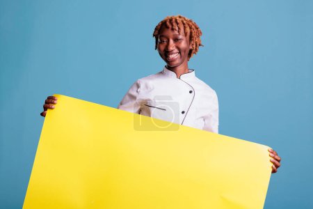 Photo for Enthusiastic kitchen helper, uniformed african american, holding a billboard, banner, advertisement. Young afro chef showing a sign. Female member of restaurant advertising. - Royalty Free Image