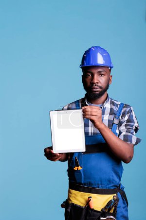 Téléchargez les photos : Vertical shot of construction worker holding digital tablet blank screen for advertising in studio shot. Serious builder dressed in coveralls showing modern electronic device against blue background. - en image libre de droit