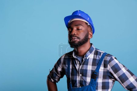 Foto de Tired exhausted construction worker in construction helmet and coveralls. African american employee with closed eyes feeling fatigued after hard work isolated on blue background. - Imagen libre de derechos