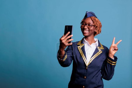 Photo for Funny female flight attendant on video call with friends during break time at work. Stewardess dressed in uniform using mobile phone while having fun chatting, studio shot. - Royalty Free Image